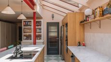 A pink kitchen with wooden cabinets, white marble countertops, an exposed red beam and white glass fronted cabinet.