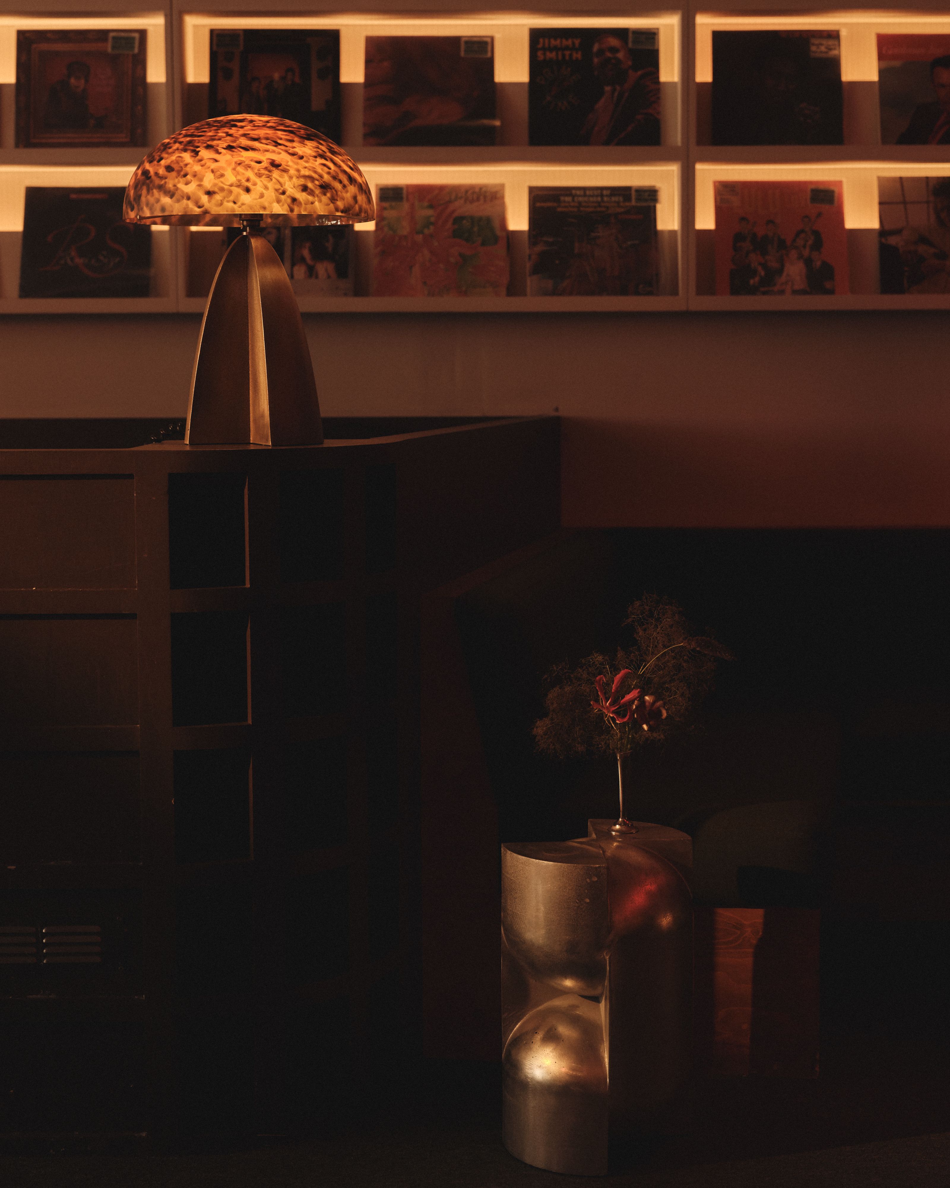 A mushroom-shaped table lamp with a chrome basis and a textured glass top sits atop a wooden cabinet in a record shop bathing in golden light alongside a chrome coffee table topped with flowers.