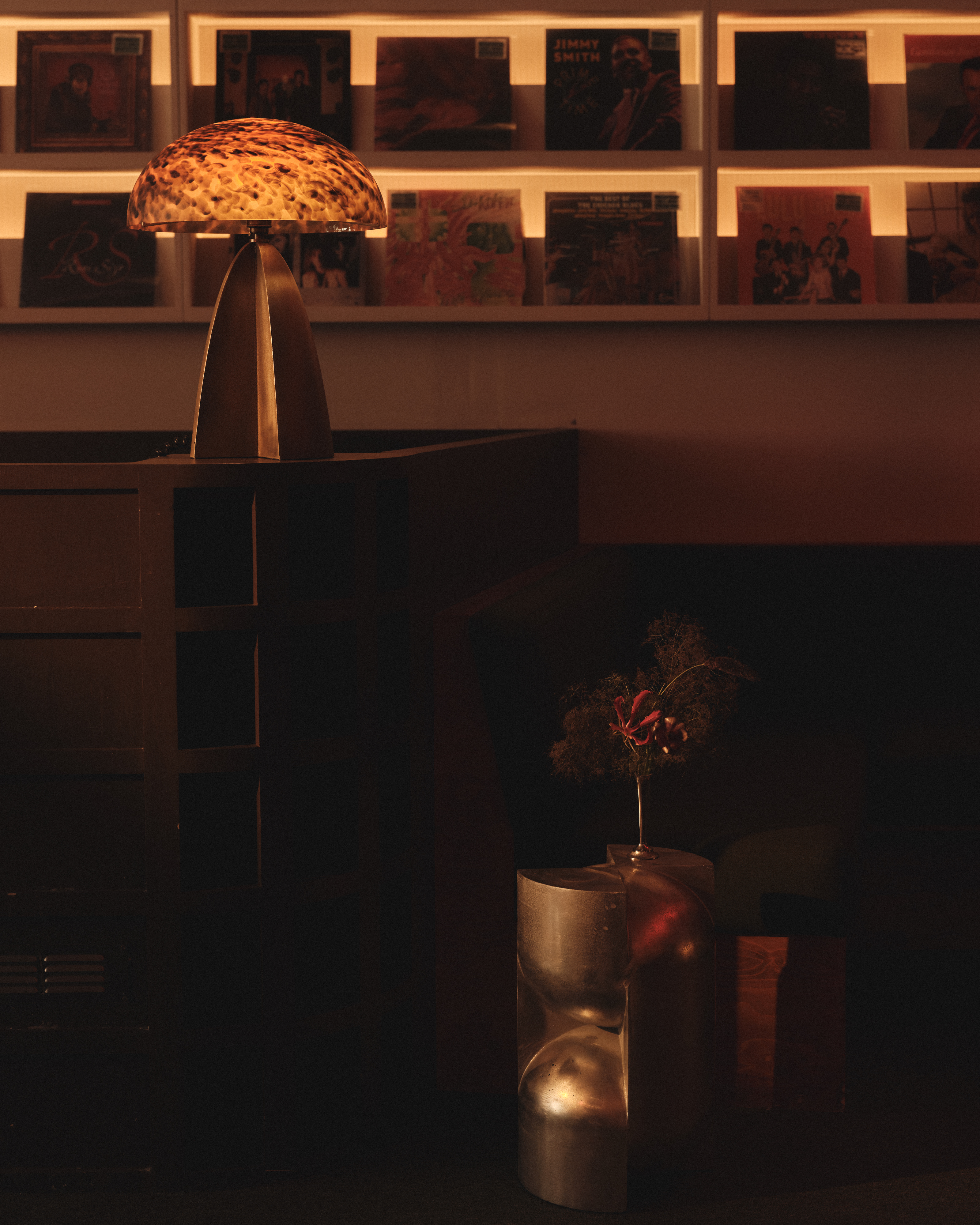 A mushroom-shaped table lamp with a chrome basis and a textured glass top sits atop a wooden cabinet in a record shop bathing in golden light alongside a chrome coffee table topped with flowers.
