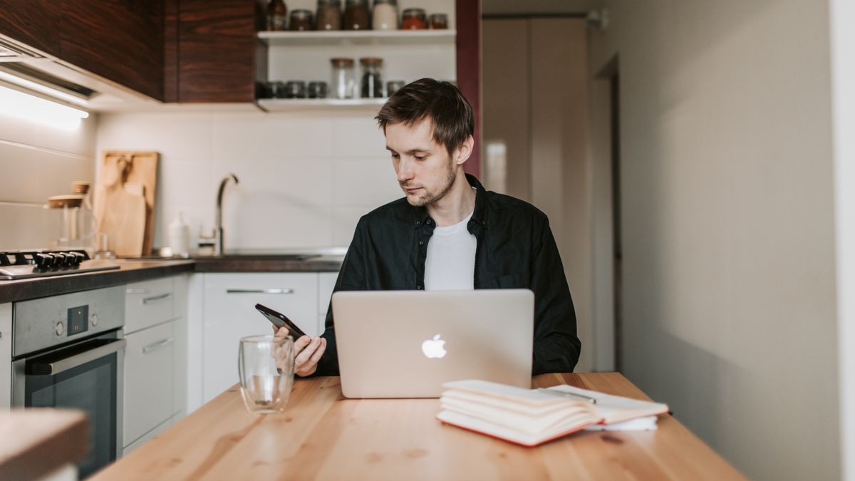 A casually-dressed man in a kitchen with a laptop, working from home.