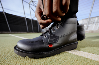 A child's hand pictured on a football pitch, fastening the laces on a pair of Kickers school shoes