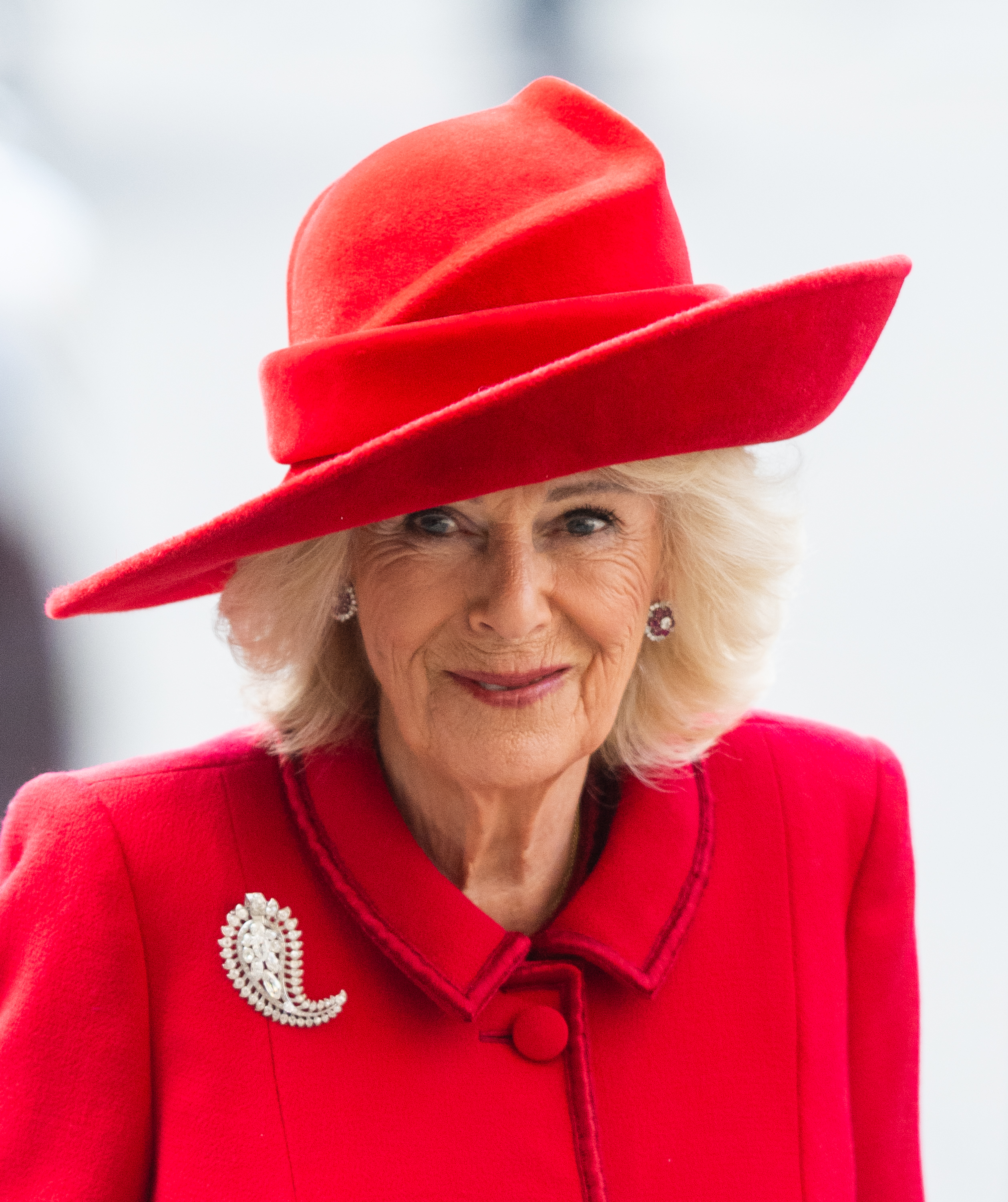 Queen Camilla wearing a red coat and hat with a diamond brooch