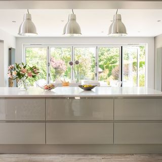 Kitchen extension with large kitchen island with neutral gloss units and light worktop and pendant lights over the top, with bifold doors in the background