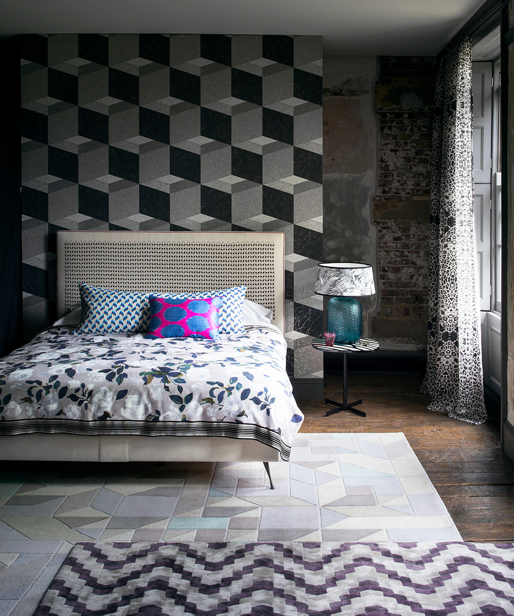 Geometric bedroom wallpaper in a dark, moody bedroom scheme with mismatched patterns including two different rug designs, floral bedlinen and spotty pillows.