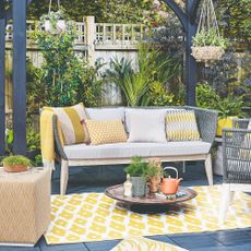 A patio with a Scandi-style outdoor 3-seater sofa with light grey seat cushions and woven dark grey back paired with ochre scatter outdoor cushions