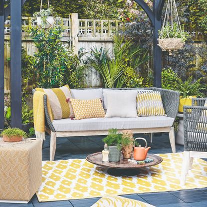 A patio with a Scandi-style outdoor 3-seater sofa with light grey seat cushions and woven dark grey back paired with ochre scatter outdoor cushions