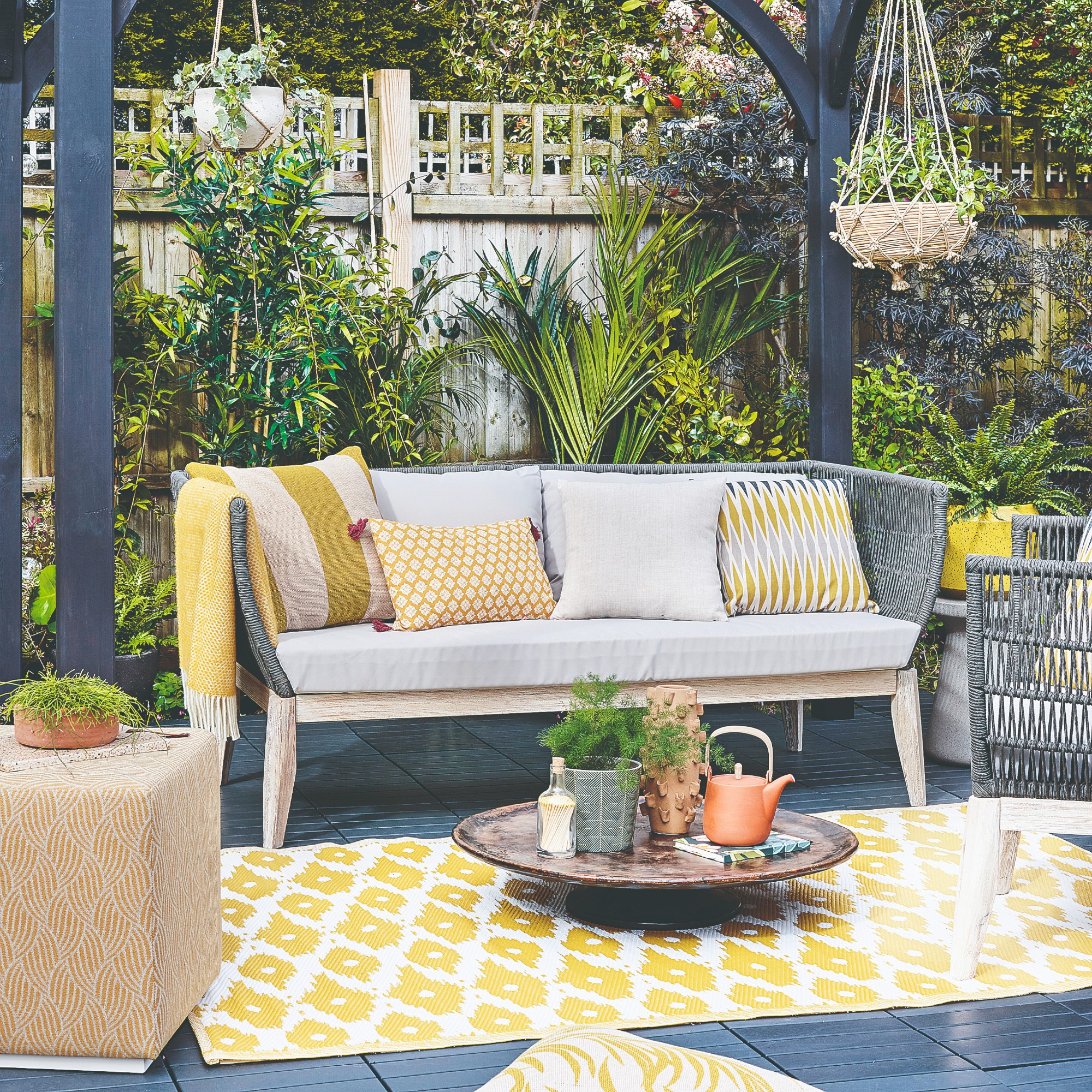 A patio with a Scandi-style outdoor 3-seater sofa with light grey seat cushions and woven dark grey back paired with ochre scatter outdoor cushions