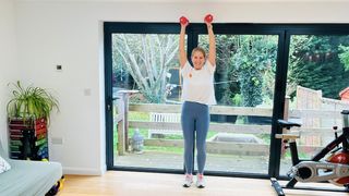 Maddy Biddulph performs an overhead press with a pair of dumbbells in her living room. She wears leggings, sneakers and a t-shirt. She is standing with her arms straight up in the air and a pair of dumbbells in her hands. Behind her we see glass sliding doors leading to a garden and an exercise bike.