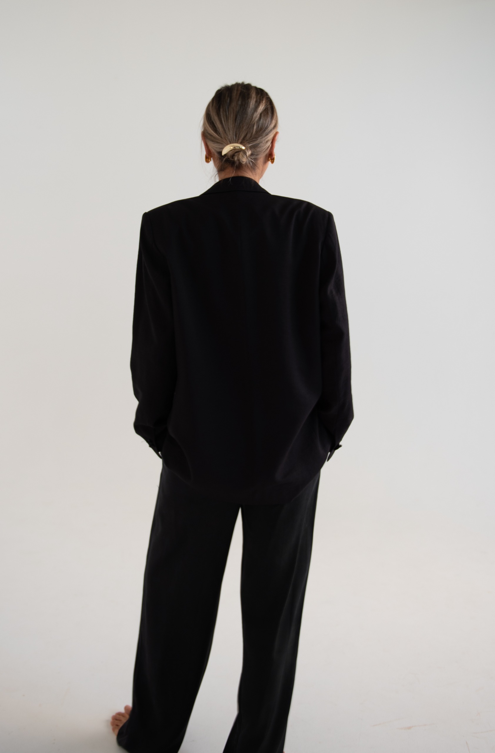 Rear view of a middle aged woman in an oversized black suit, standing against a plain white studio background.