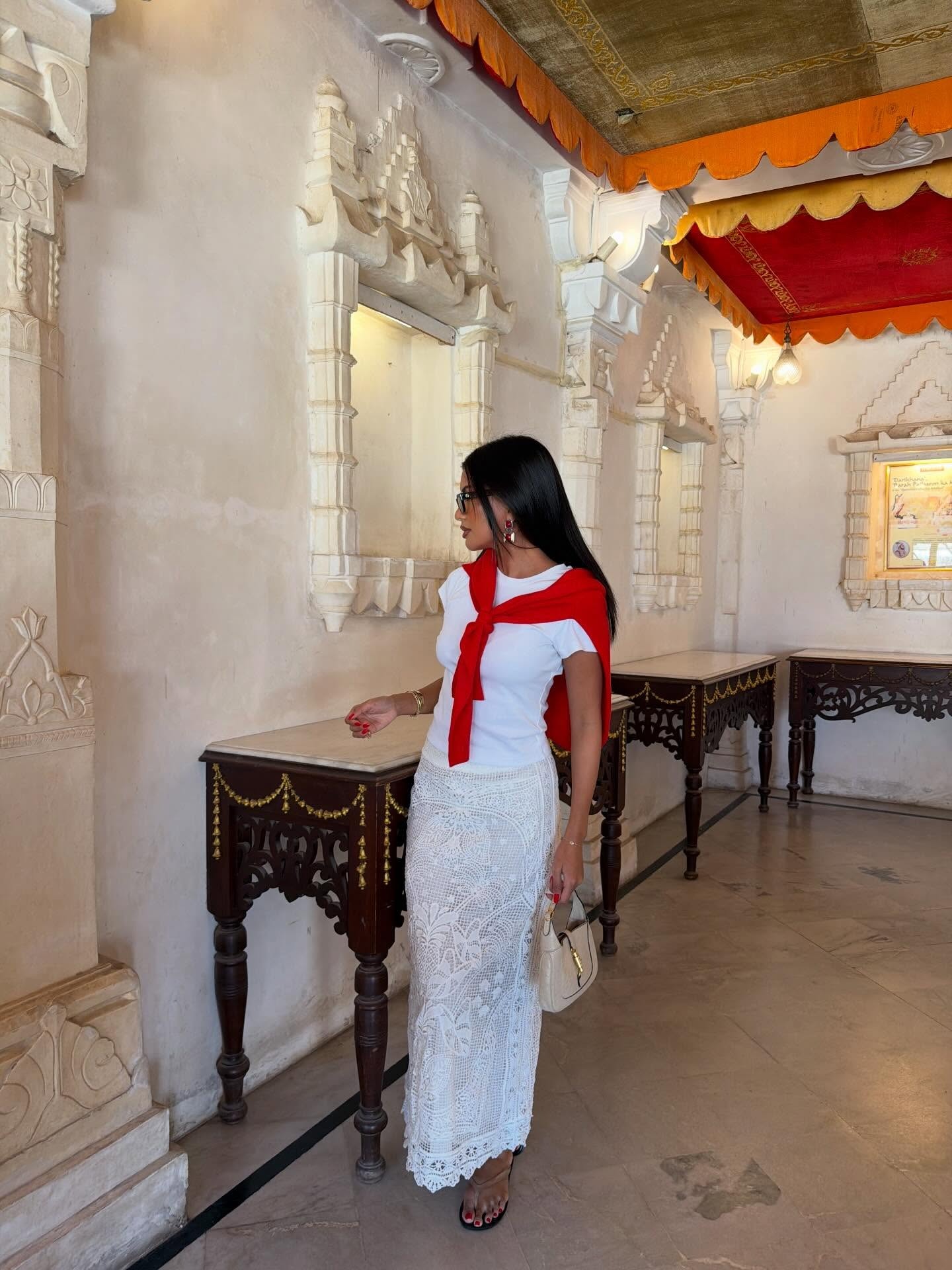 Fashion person Pia Shah wears a winter sun outfit. She wears a white tee shirt, lace maxi skirt and red jumper. She finishes the look with clear jelly sandals. This image was recently taken from her Instagram.