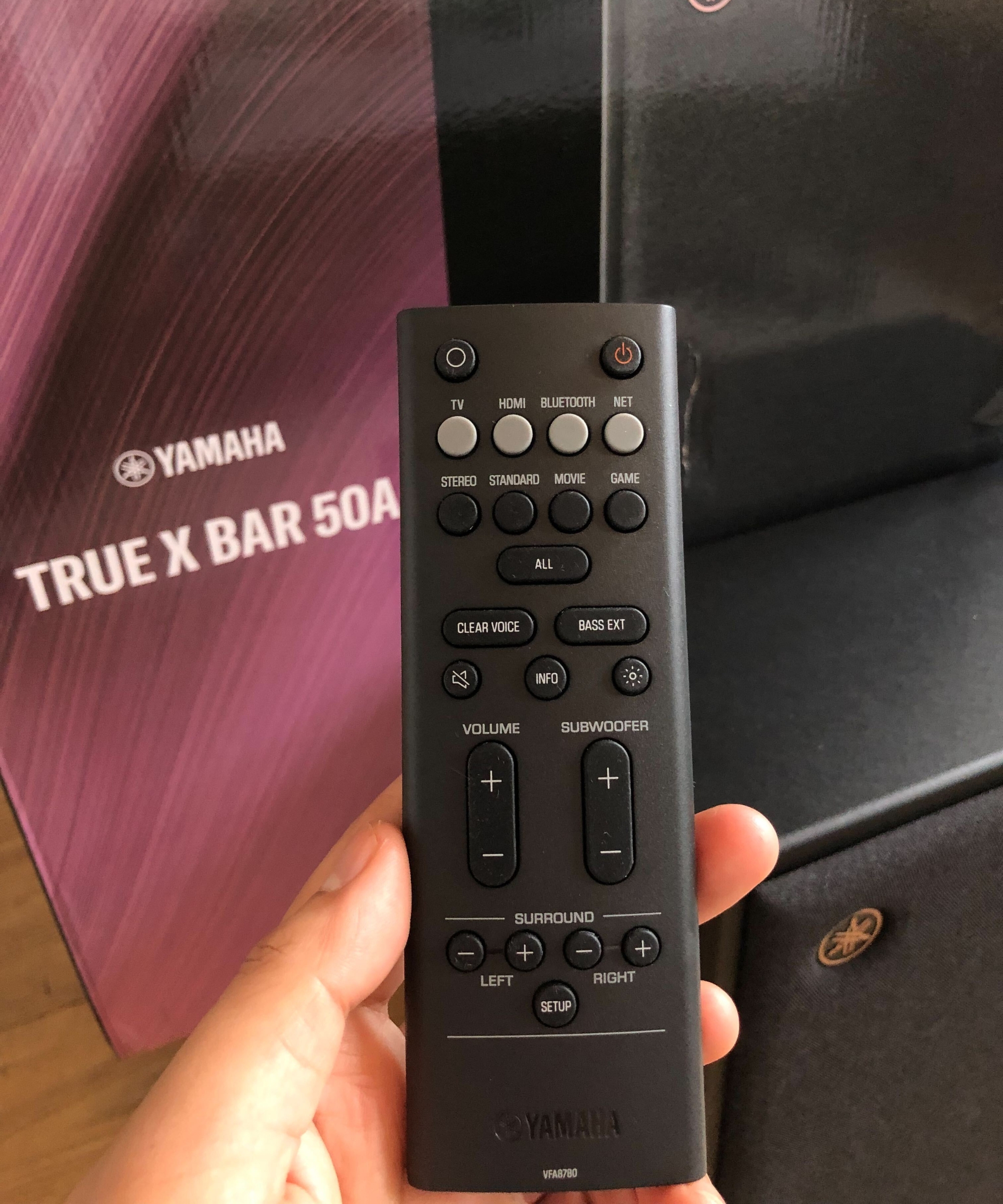 Hand holding the Yamaha True X Bar 50A remote in front of the box and subwoofer.