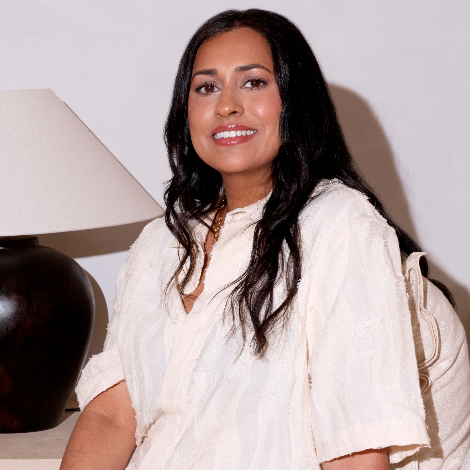 A woman with wavy black hair in a white blouse with three quarter sleeves on a sofa beside a black and white lamp