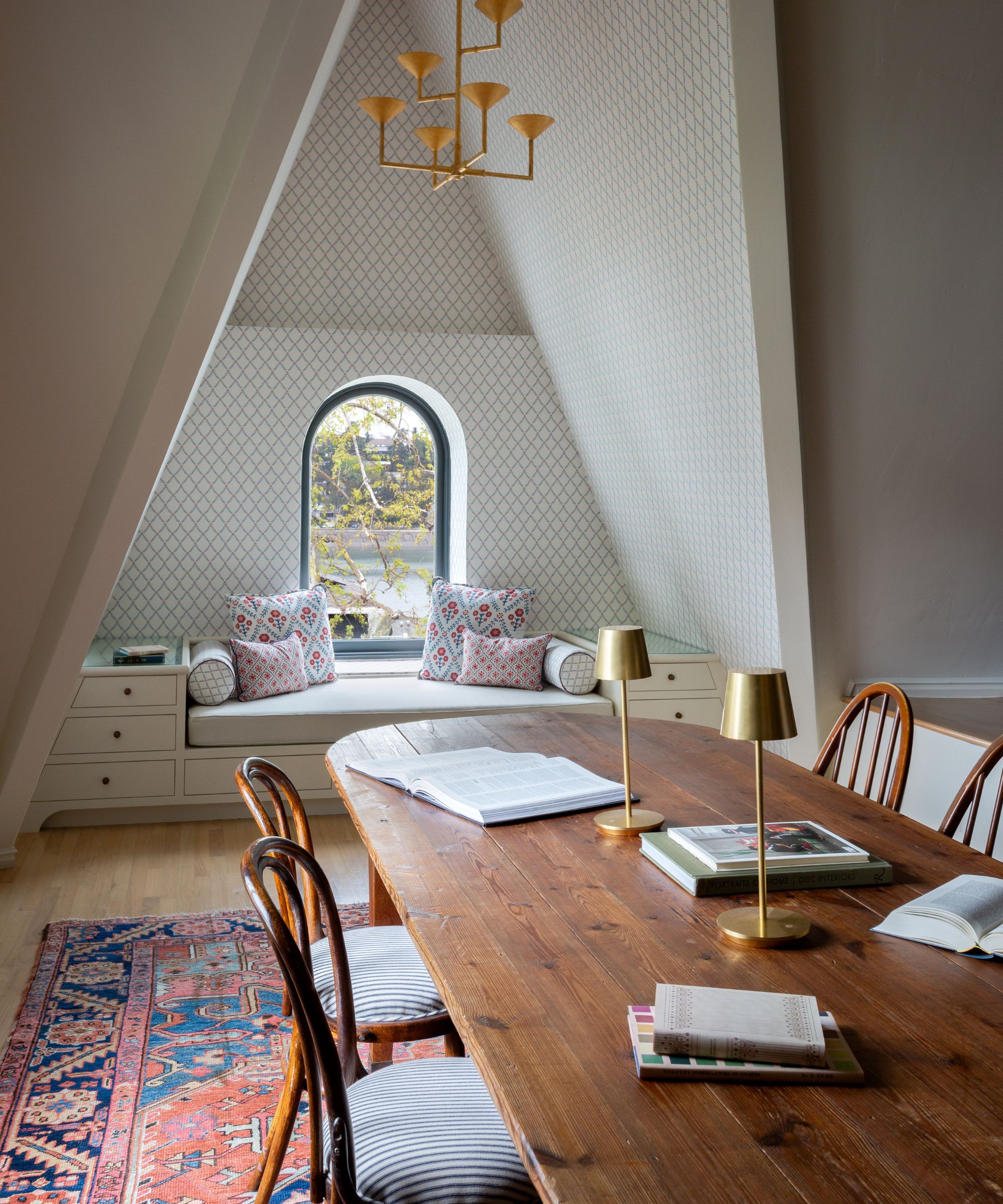 a vaulted ceiling upstairs family room with wallpapered window nook and custom day bed, with a large dining table and chairs with a red and blue vintage rug