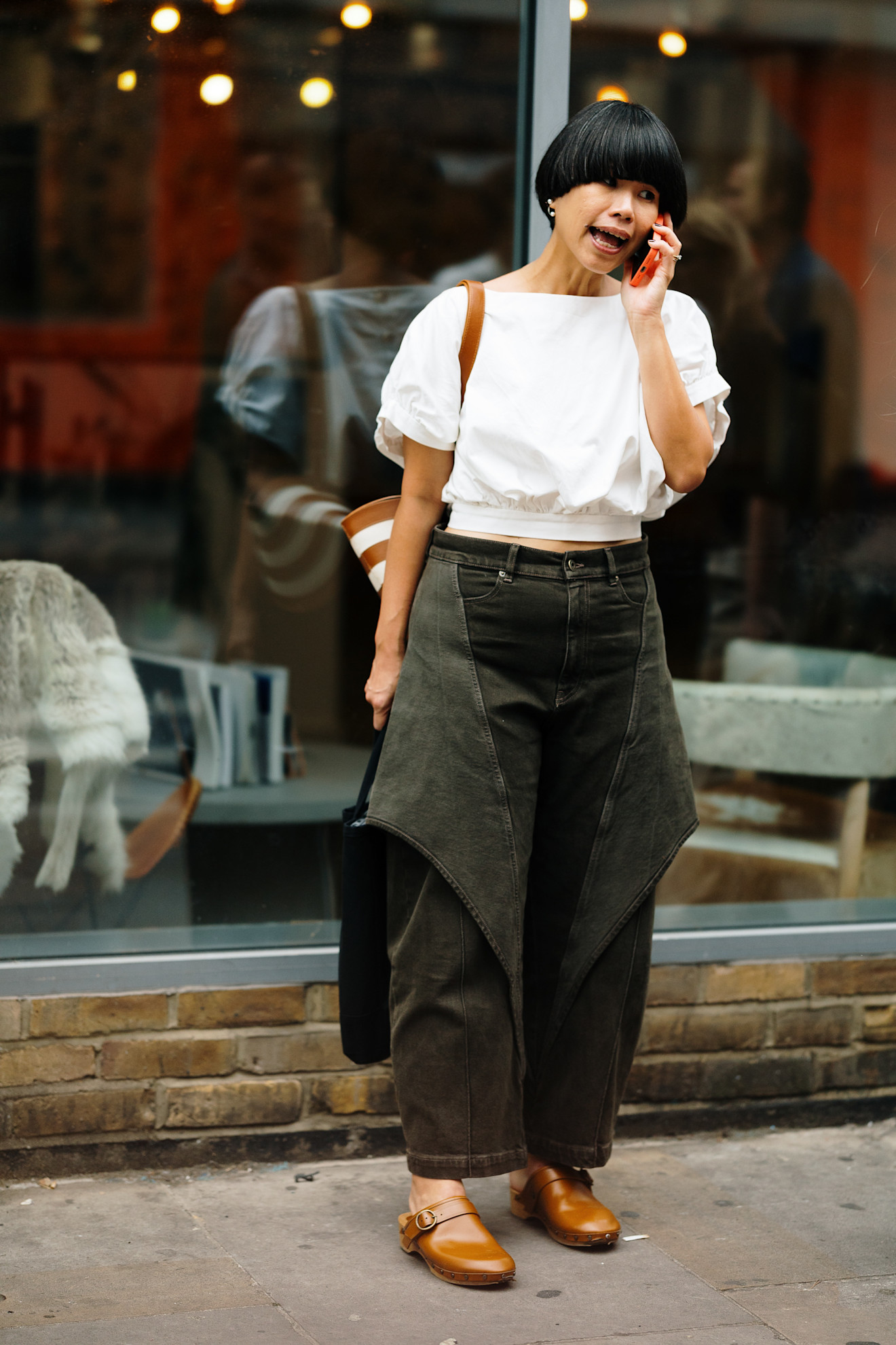 a woman with a dark brown bowl cut talking on the phone wearing dark jeans and a white blouse