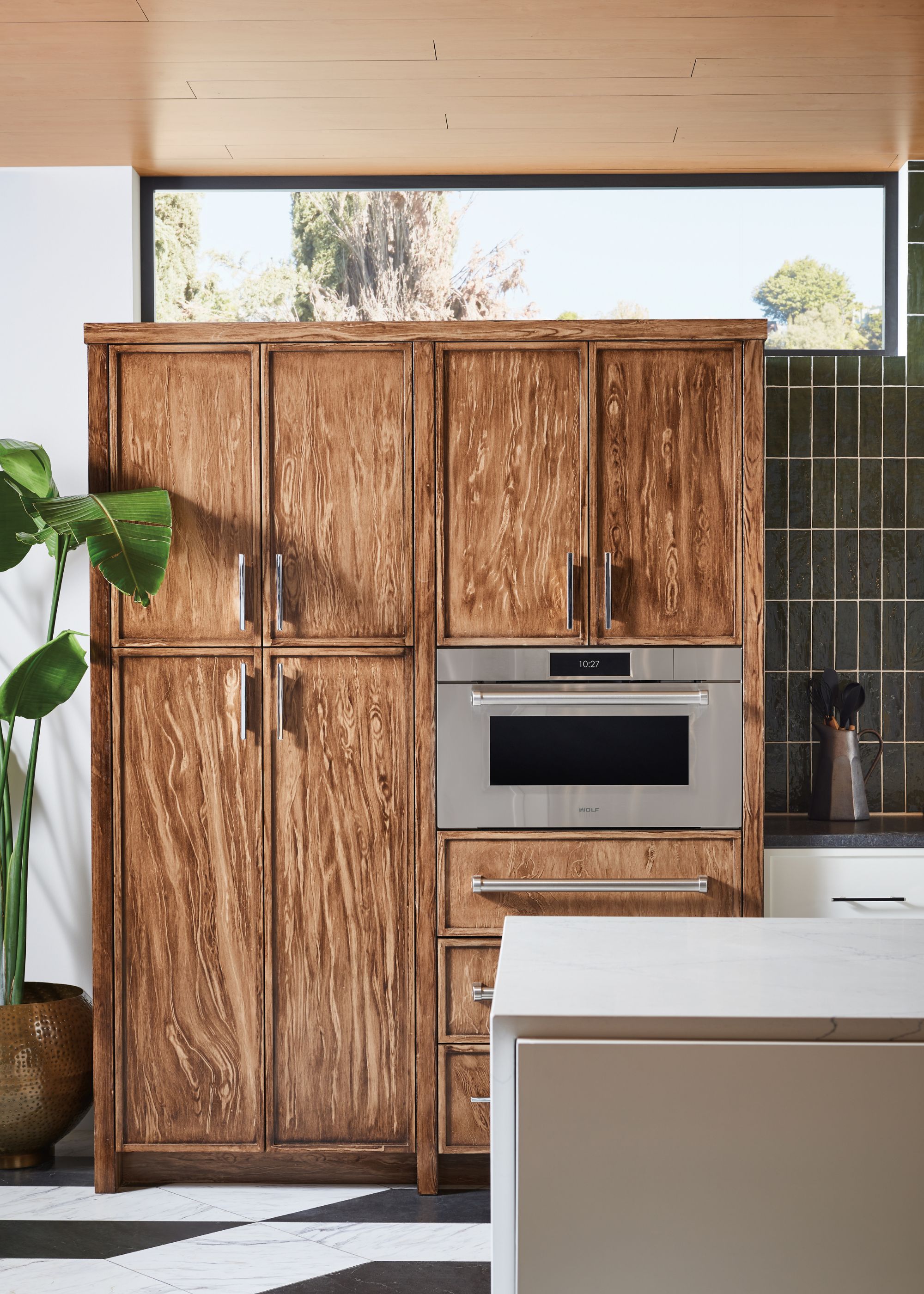 A kitchen with wooden cabinets and a proving drawer placed on one side as well as a corner view of a large plant in a pot as well as a marbled kitchen island