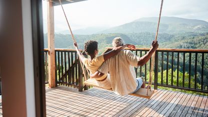 An older couple sit on a porch swing together facing a view of a valley.