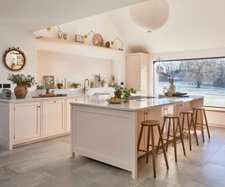 A spacious kitchen with soft pink cabinetry and a large island topped with white marble. Four wooden barstools with round seats and tapered legs are aligned along the island. A brass faucet sits over a farmhouse-style sink, with small potted plants and decorative items on the countertop