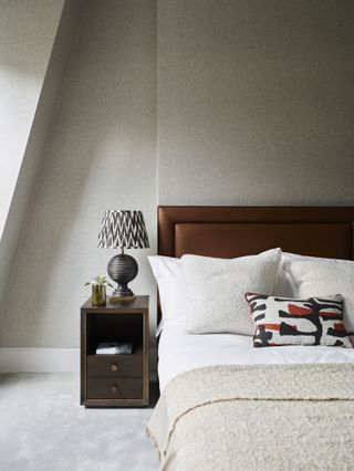 A bedroom with a wooden bed frame, beige sheets, and cream carpet and white baseboards