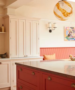 The corner of a red kitchen island with a dark brown countertop, and pink cabinets behind it and a orange and white glass pendant light above it