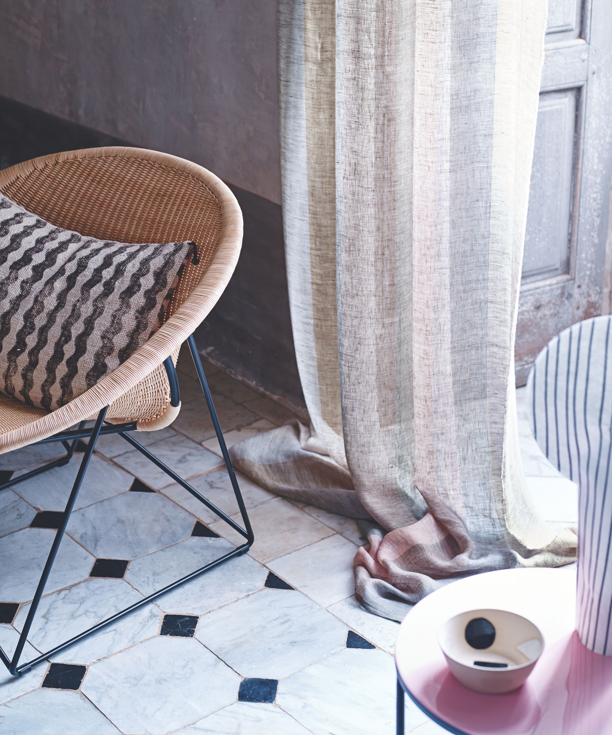 Sunny open wooden door with sheer linen curtain, black and navy tiled marble floor and rattan chair with a black and white striped cushion. In the foreground to the right is a pink circular table with patterned bowl and patterned vase.