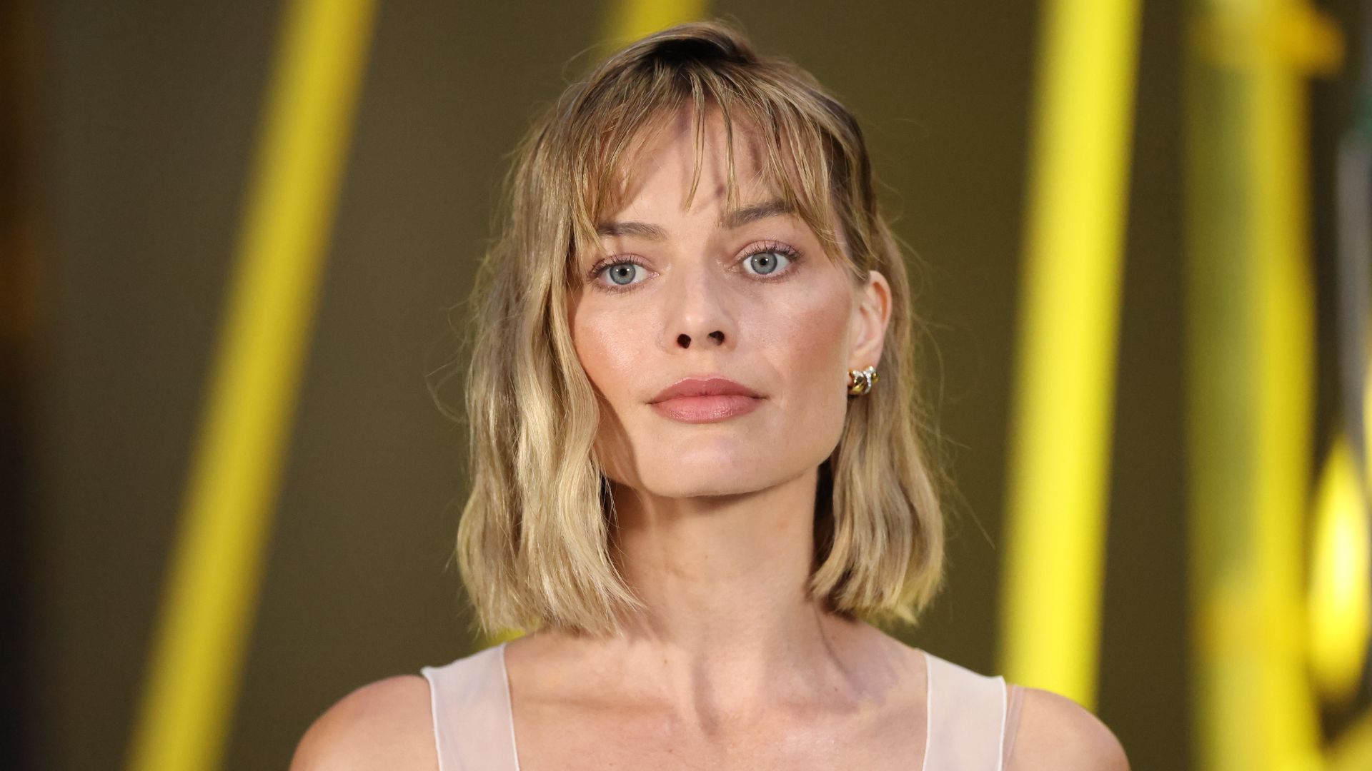 Margot Robbie pictured with a bob and fringe haircut wearing a white vest top against a yellow backdrop