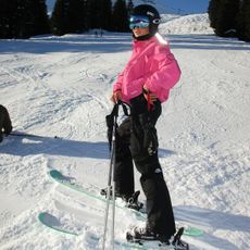 Emili wears a pink puffer jacket, black ski pants, and blue goggles.