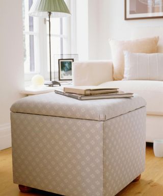 A gray storage ottoman cube with a diamond pattern with two books on top, with white walls in the background, a green lamp, and a white bed with shiny brown and silver throw pillows on it