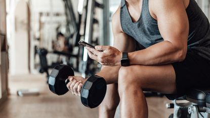 A muscular Millennial man works with a barbell in a gym while looking at his cell phone.