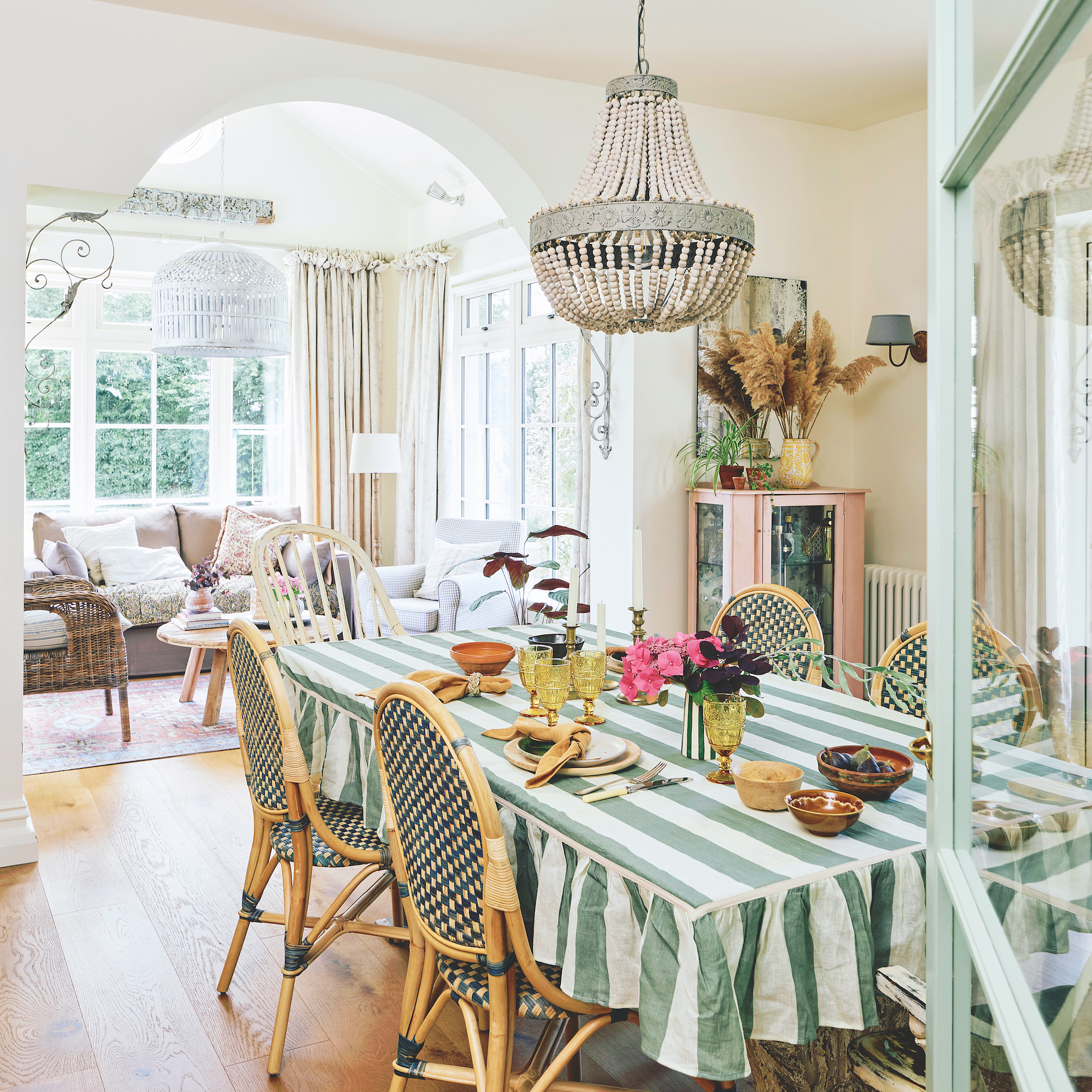 dining room table covered in white and green stripe cloth with green woven rattan chairs and bead chandelier above