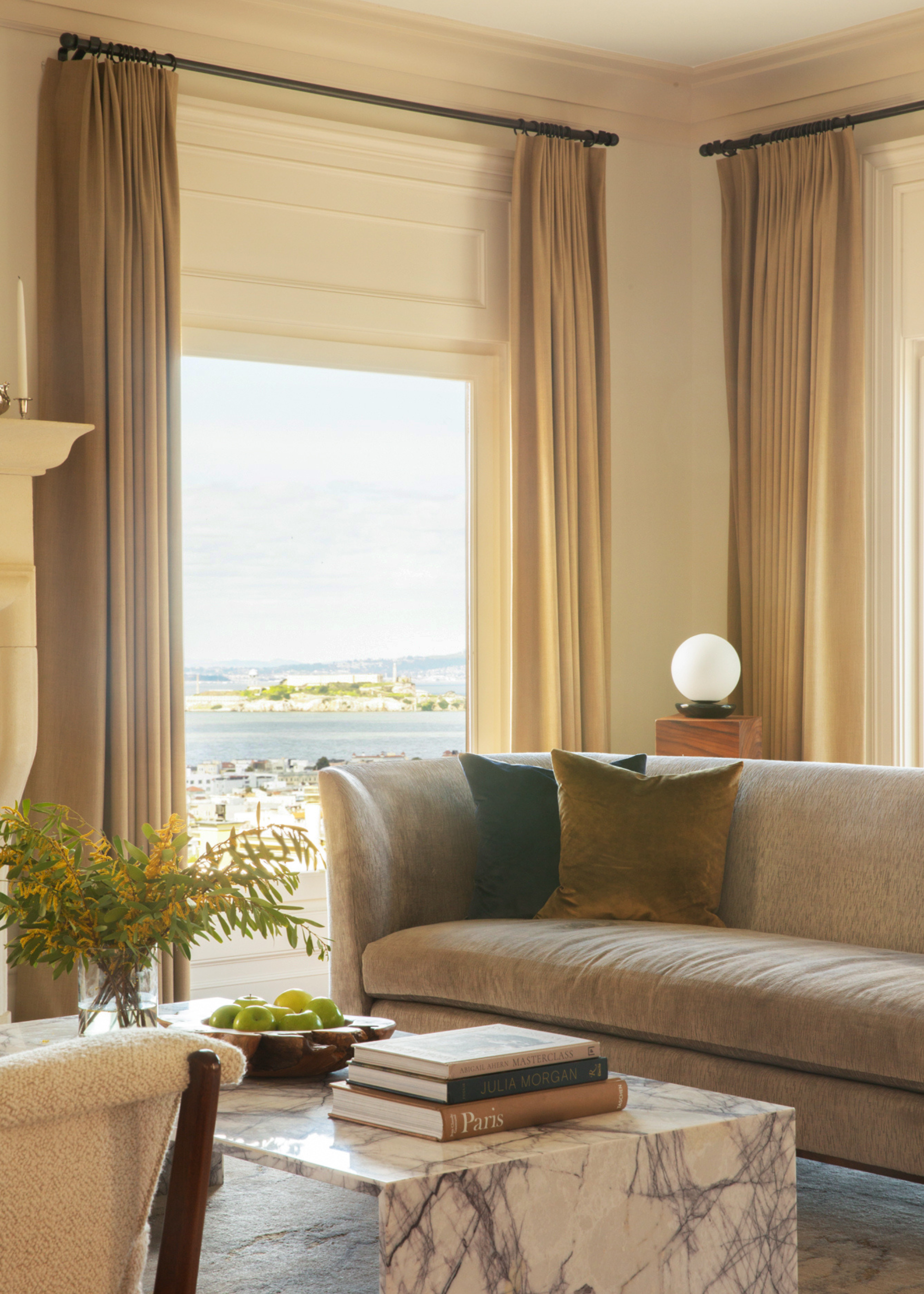 A bright living room with large windows, a gray couch, square cushions, a marble coffee table with a bowl of fruit, a vase of stems, and a stack of books
