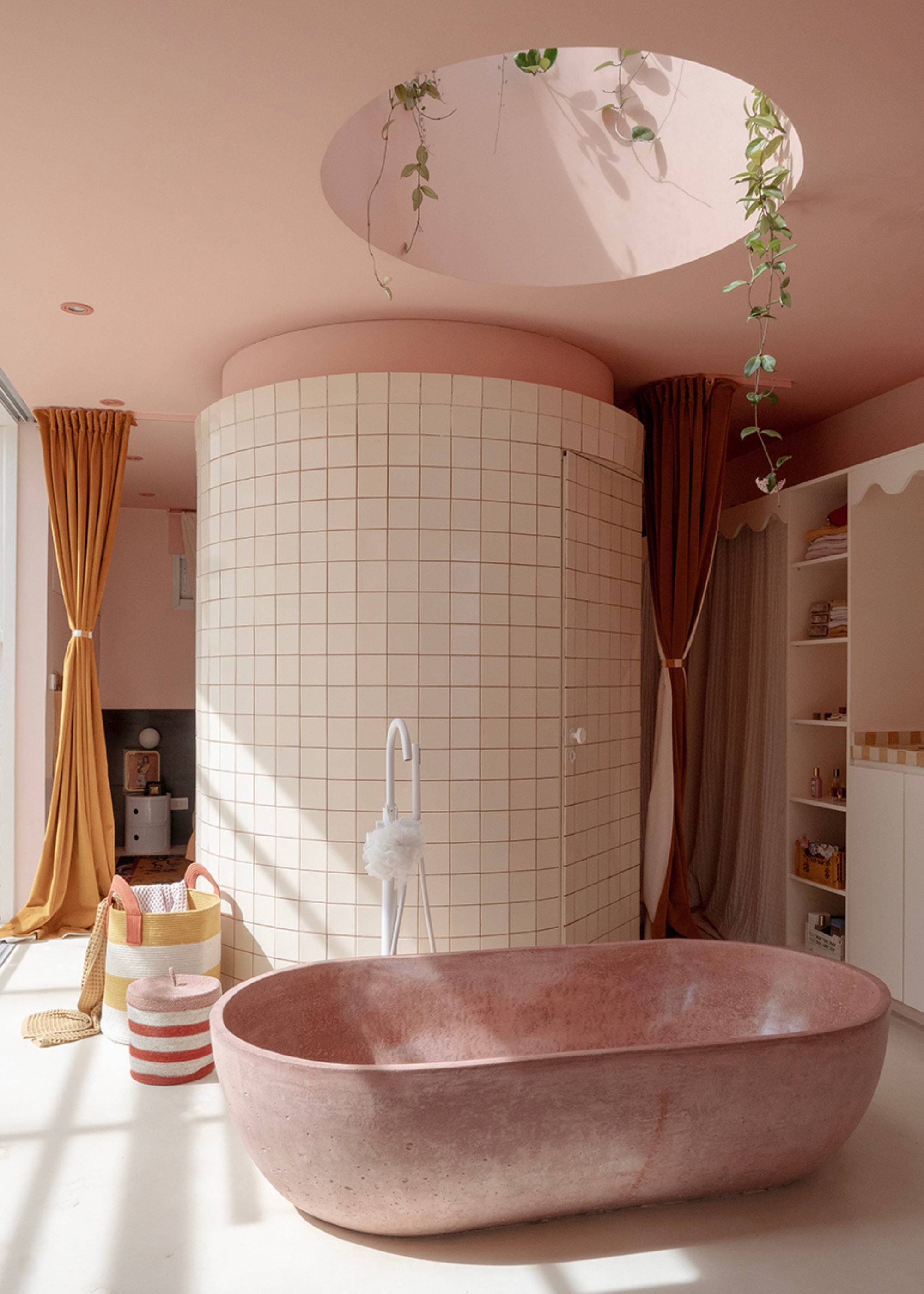 A pale pink bathroom with a skylight, a pink tub, a white tap, a striped hamper, a sound tiled center wall with a door to a bathroom, and a shelving unit