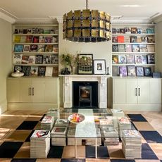 Living room with built in magazine display shelving above cupboards on either side of marble fireplace in front of glass coffee table held up by piles of magazines