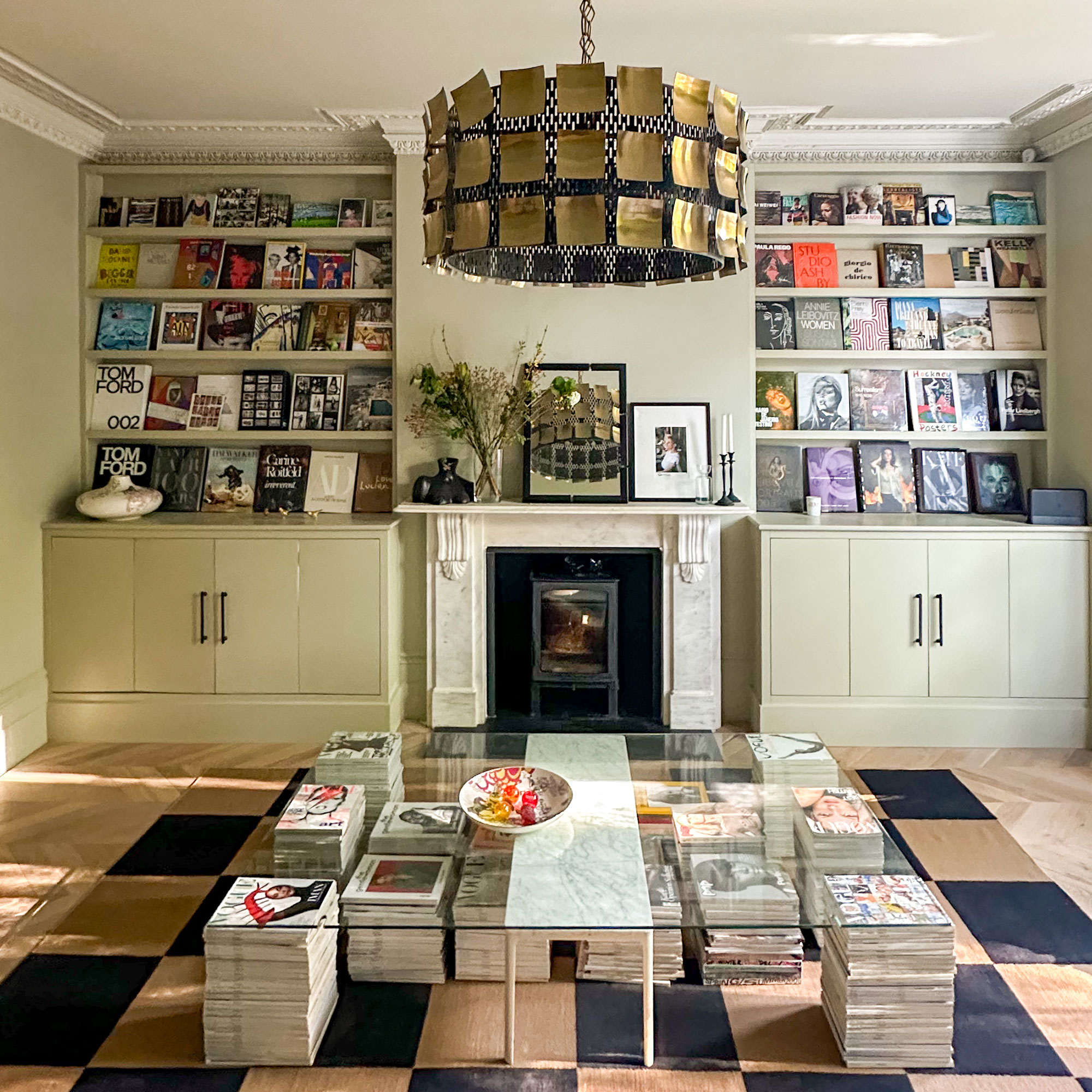 Living room with built in magazine display shelving above cupboards on either side of marble fireplace in front of glass coffee table held up by piles of magazines
