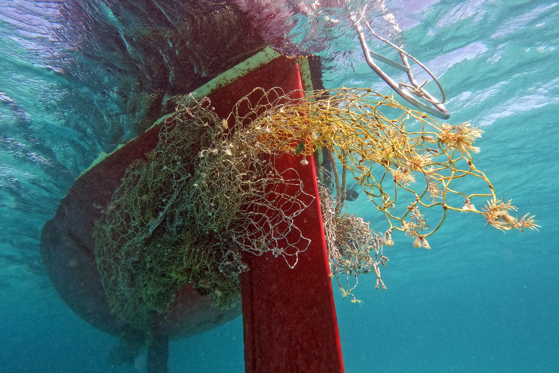 An underwater view of a red boat hull and keel entangled in a large, messy mass of discarded yellow and brown fishing nets in clear blue water.