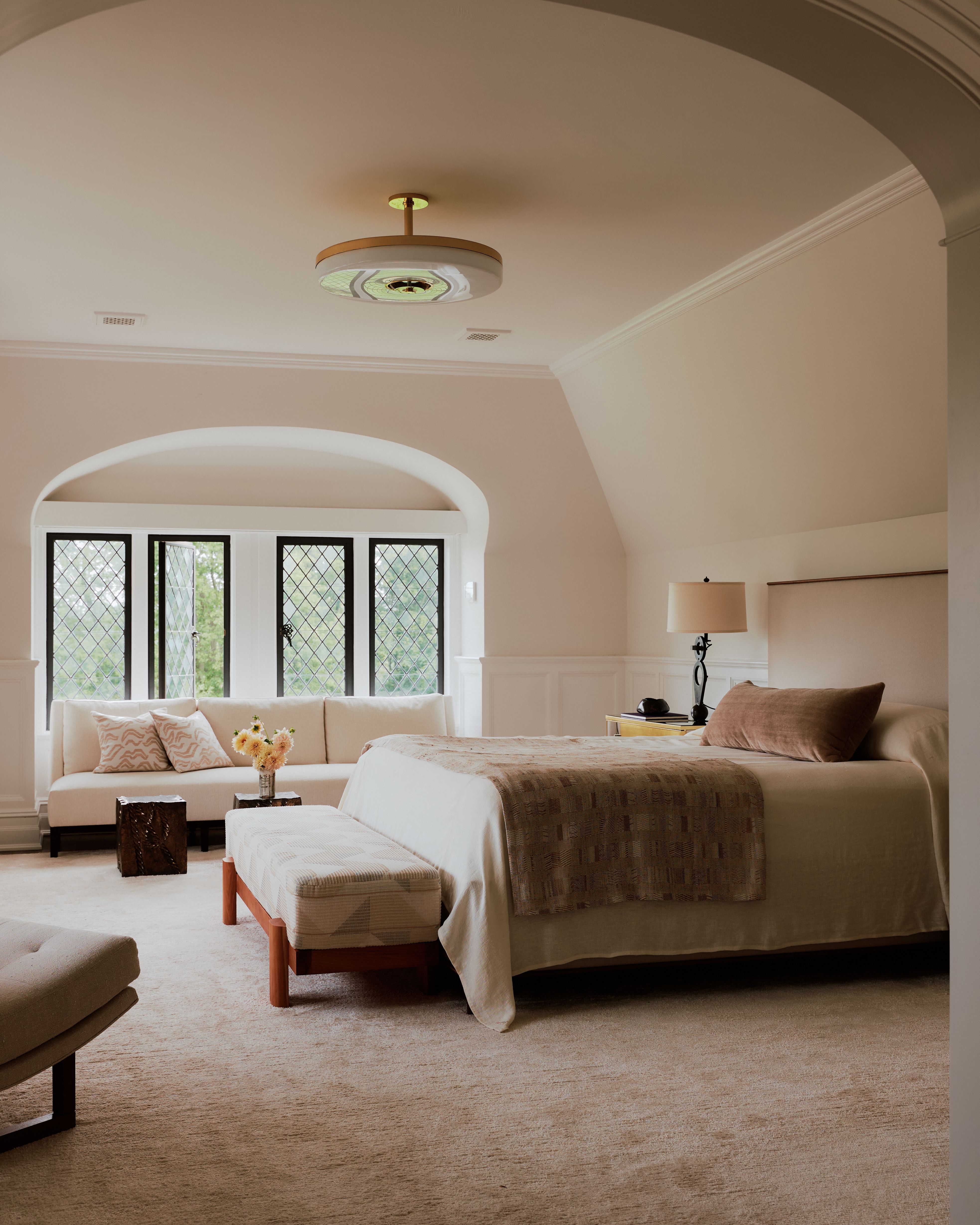 A champagne-tinted bedroom with an arched entrance, fuzzy beige carpet, and elegant but linear decor, features a roun pendant lamp in glass and brass.