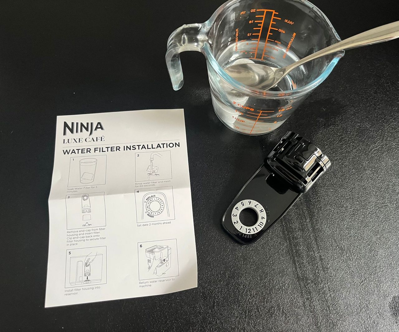 A water filter soaking in a glass jug being held down with a spoon, with an instruction manual and filter cage beside it.