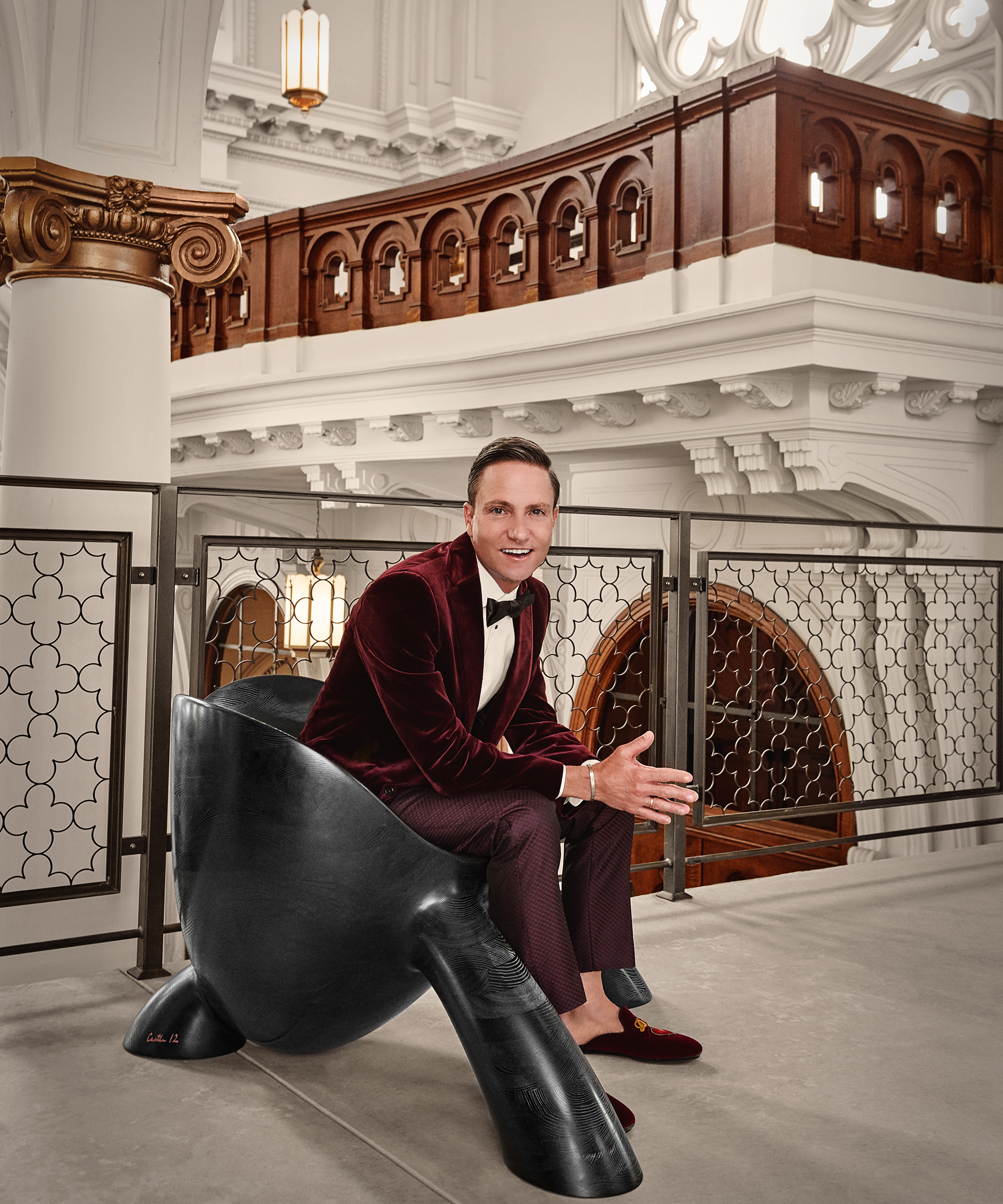 Ken Fulk in a red smoking jacket sat on a modern black chair smiling to camera