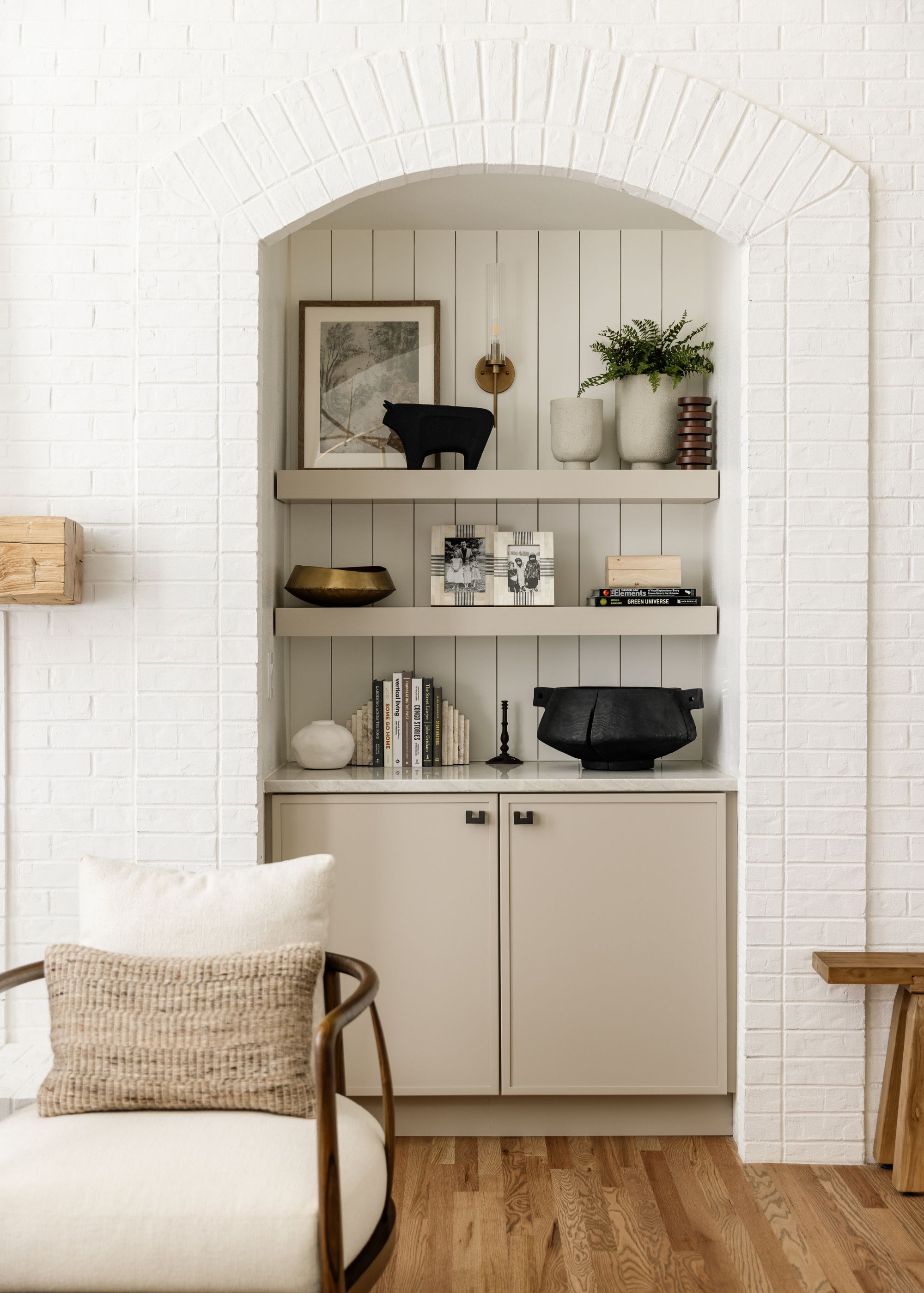 A living room with a white brick wall with alcove shelving and white paneled walls at the back. Wooden floors and a white armchair.