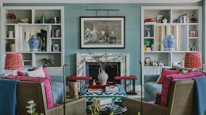 Living room with blue grasscloth walls, built-in bookshelves, marble fireplace with painting above it, pink armchairs with red lamps on either side, and two blue sofas.