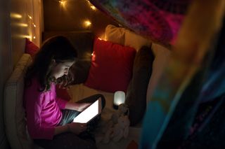 A young girl using an iPad whilst sitting in a den in her bedroom with cushion and blankets, it's dimly lit with a small lamp.
