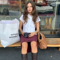 A woman sitting on a bench wearing a white button-up blouse, a red skirt, and brown knee-high boots. 