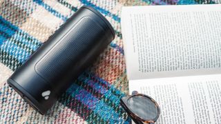A PINE Beat speaker in black, lying on a light colored tartan fabric with an open book and a pair of sunglasses beside it