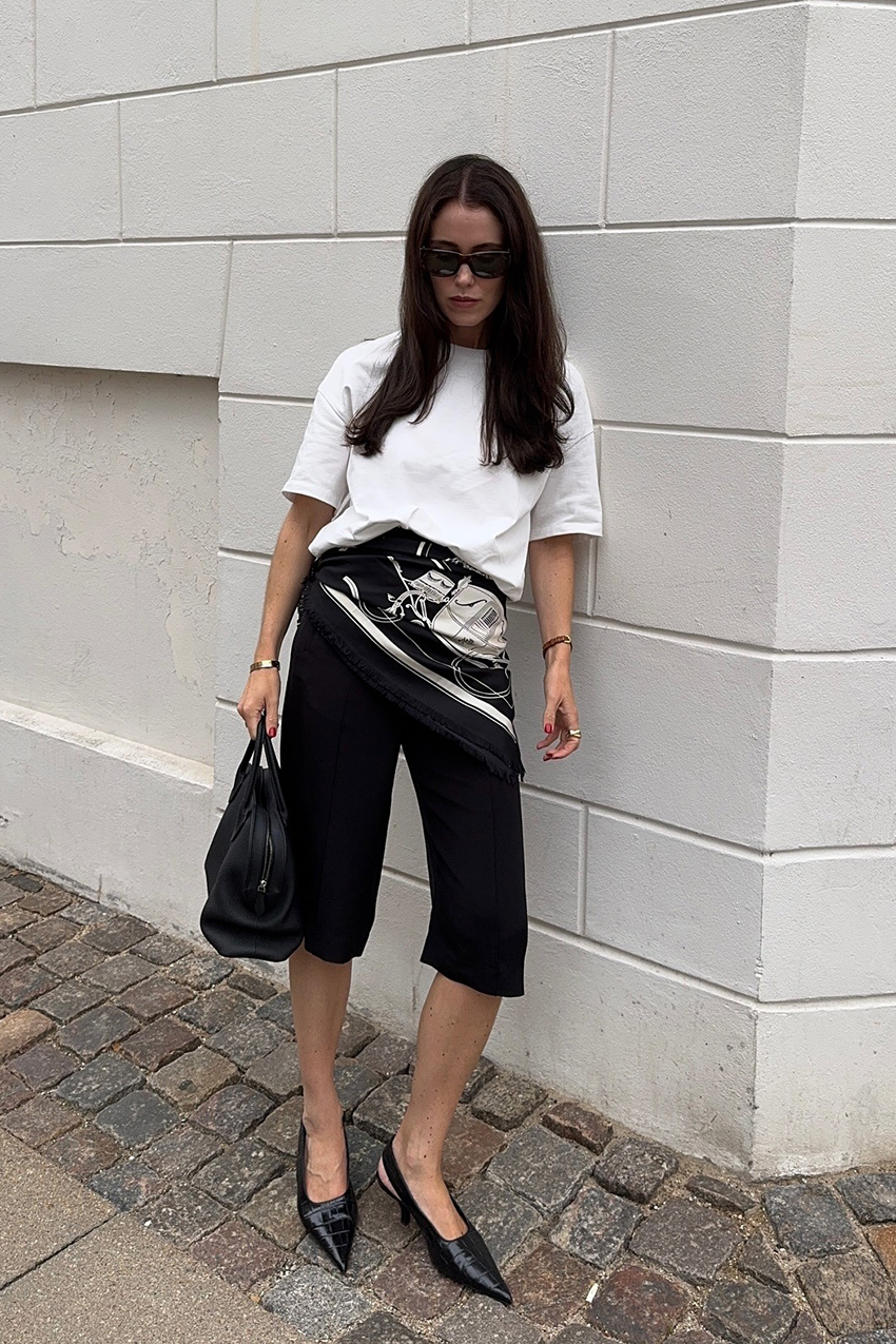 a woman wearing a white tee, black culottes, black pointed shoes, and a black leather bag