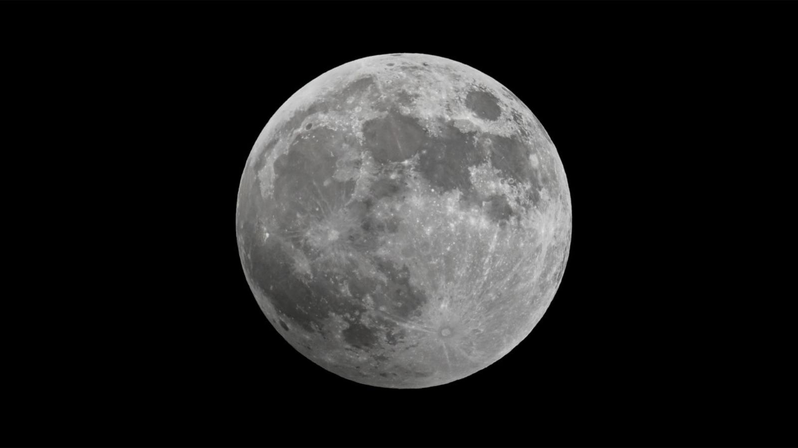 An image of a full moon suspended against a black sky. Dark patches known as lunar mare are visible on the lunar surface, which represent basaltic plains left behind by ancient lava flows.