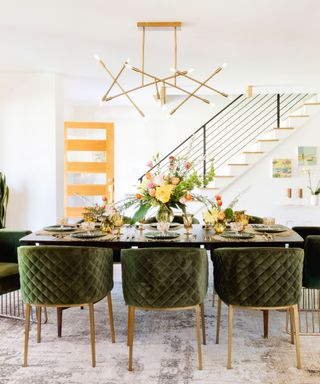 A mid-century modern dining room with a glass dining table with colorful flowers and tableware, eight dark velvet chairs tucked under it, a gold geometric chandelier, and a white staircase and a light wooden door