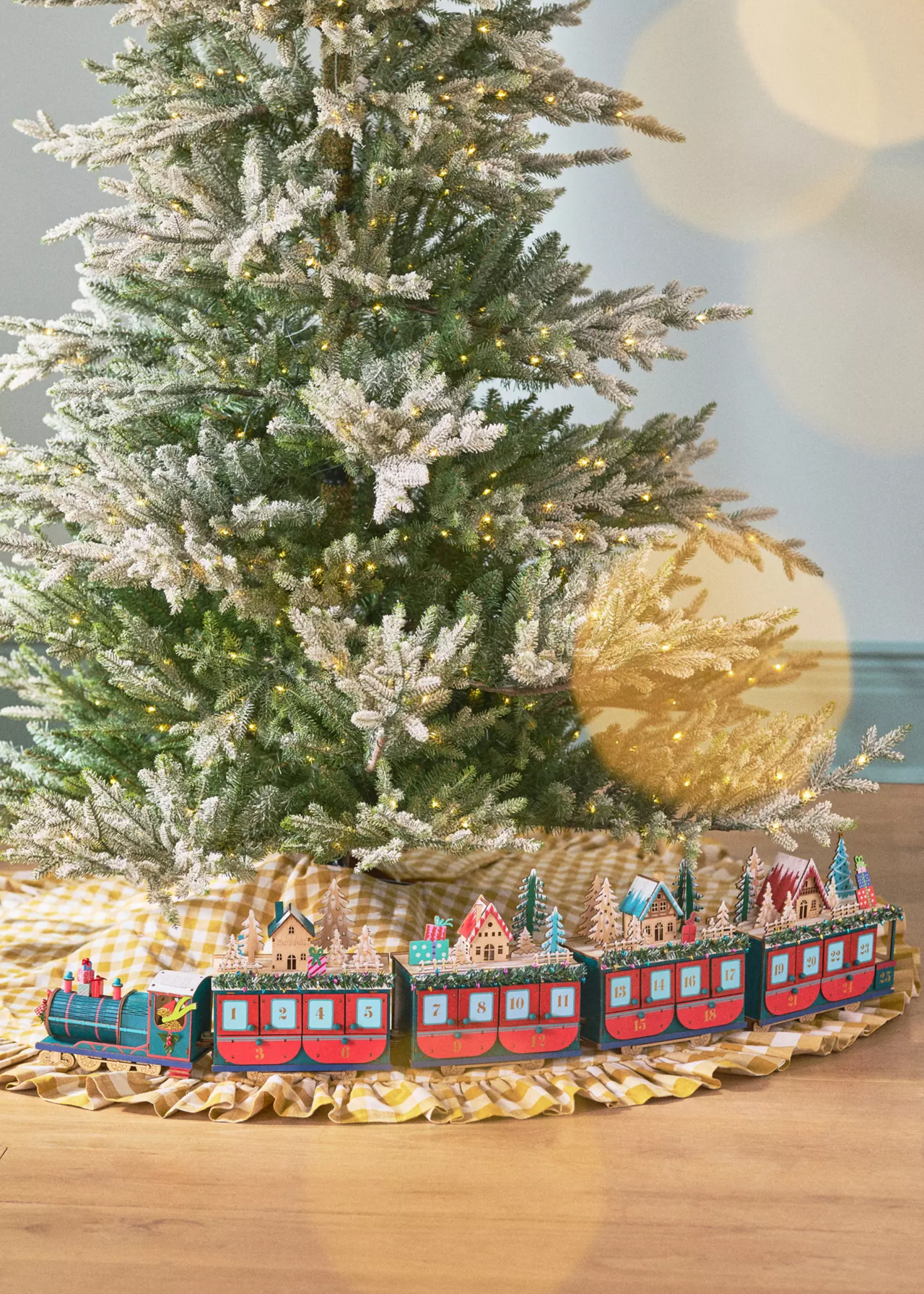the anthropologie train advent calendar placed round the gingham skirt of a frosted christmas tree, with red carriages with trees and christmas houses on top and a blue front carriage