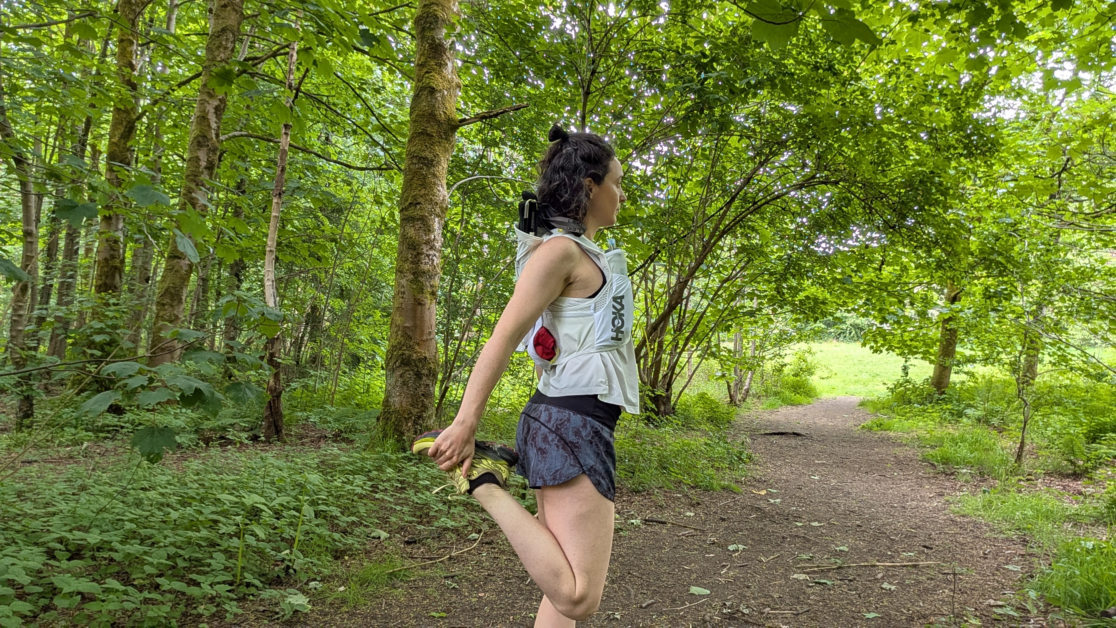 A woman trail runner doing a quad stretch in the forest wearing a white Hoka hydration vest