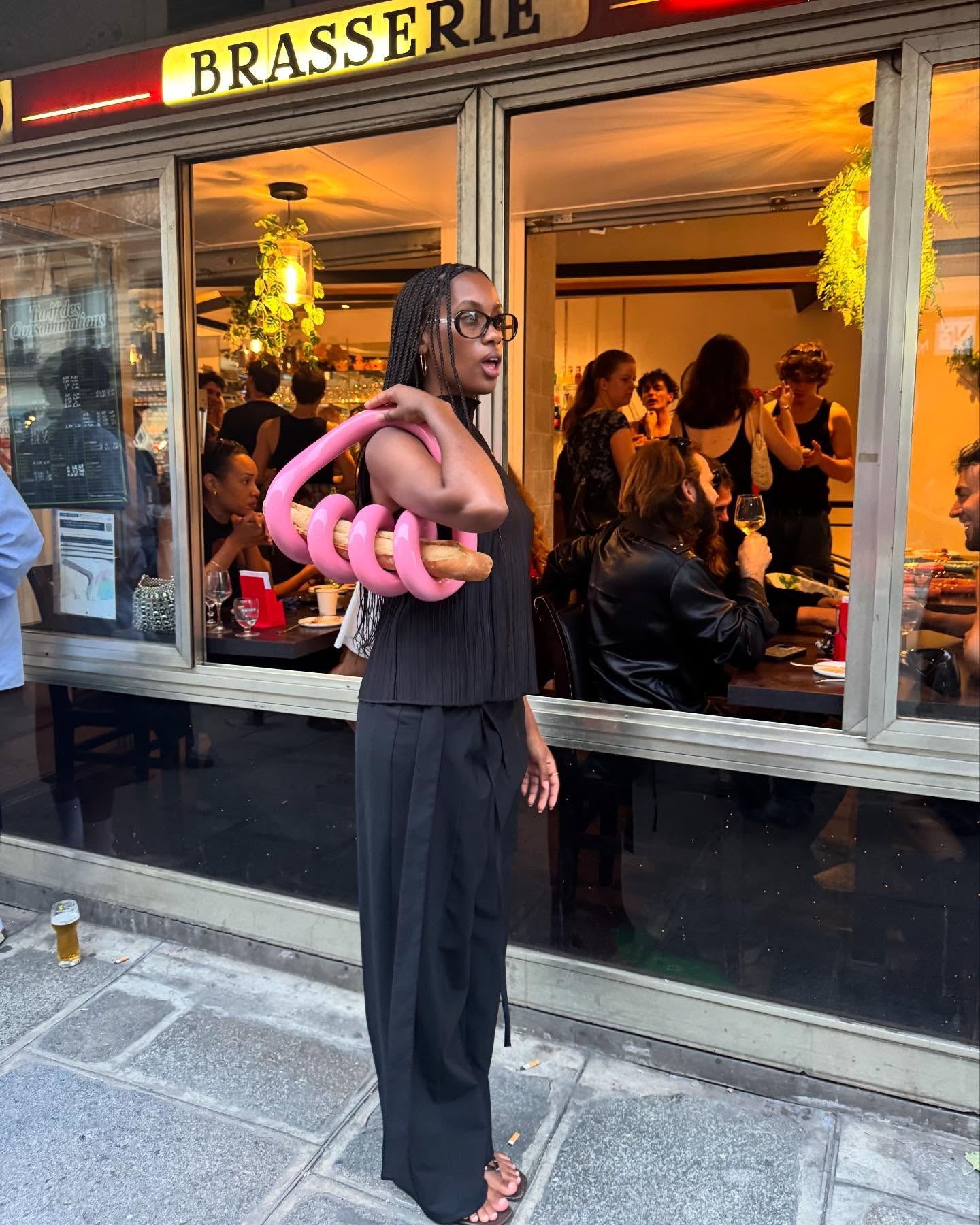 A woman wearing a black pleated top, black long pants with a pink baguette bag
