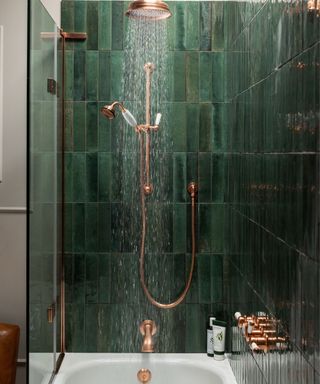 A small bathroom shower area with dark green tiles, a copper shower head and faucet, and a white bath tub with glass to the left of it