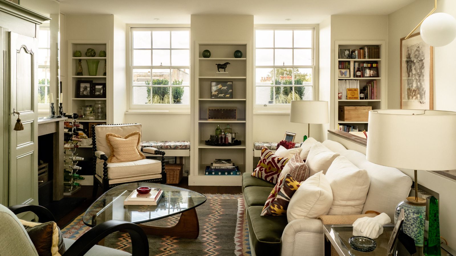 Living room with warm off-white walls, built-in shelving, layered textiles, a cream sofa, and natural light streaming through large sash windows.